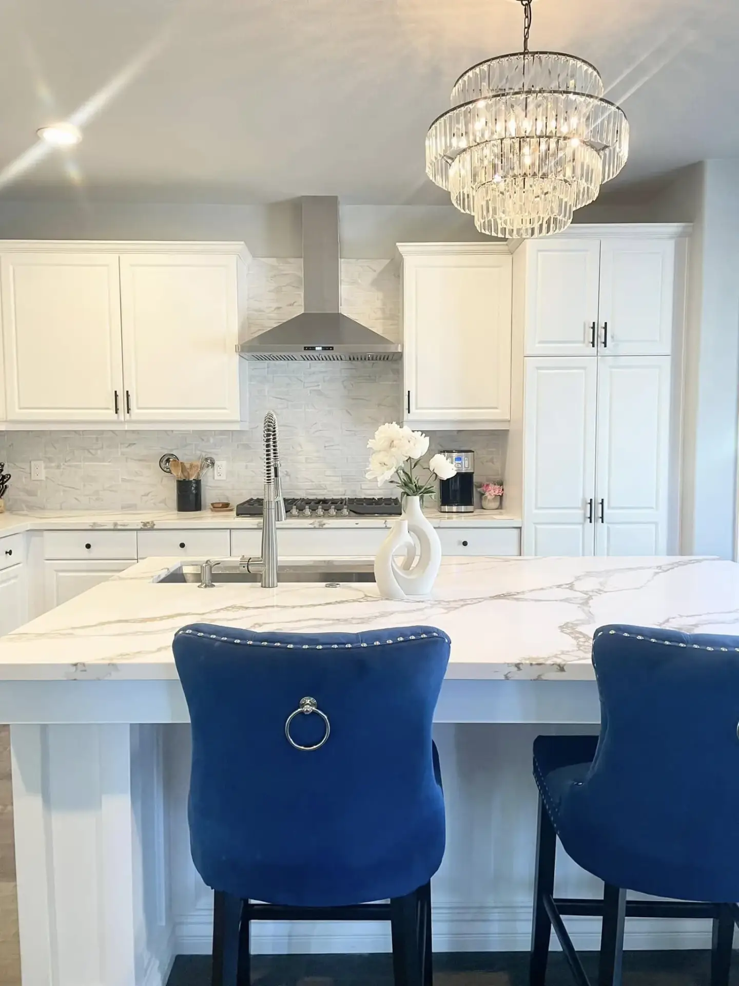 Kitchen with white cabinets, a marble-veined island and blue velvet barstools under a crystal chandelier