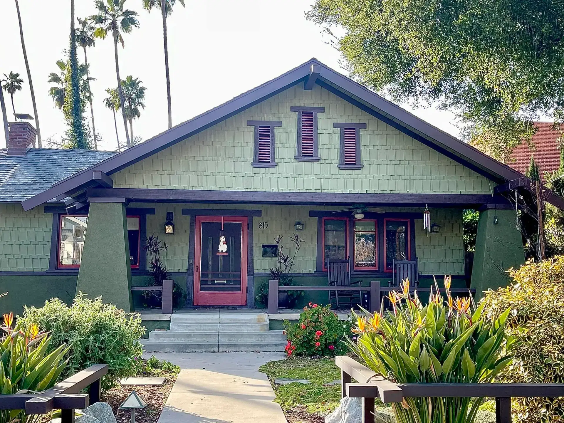 Sage green Craftsman bungalow with plum trim and a coral front door, framed by tall palms