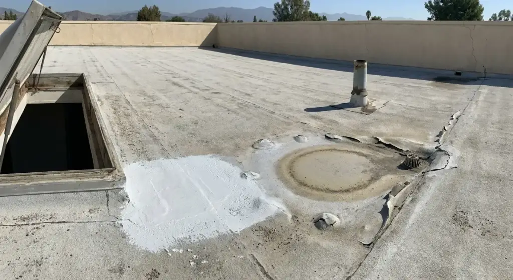 Low-slope roof showing a white coating patch beside ponding stains, blisters, and peeling near a roof drain