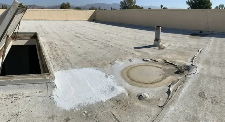 Low-slope roof showing a white coating patch beside ponding stains, blisters, and peeling near a roof drain